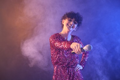 Photo of Beautiful young woman with microphone on color background in neon lights and smoke