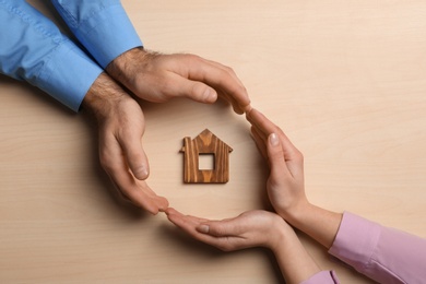 Photo of Couple holding hands near figure of house on wooden background, top view. Home insurance