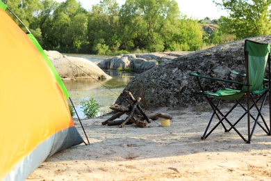 Photo of Modern camping tent with chair and firewood on riverbank