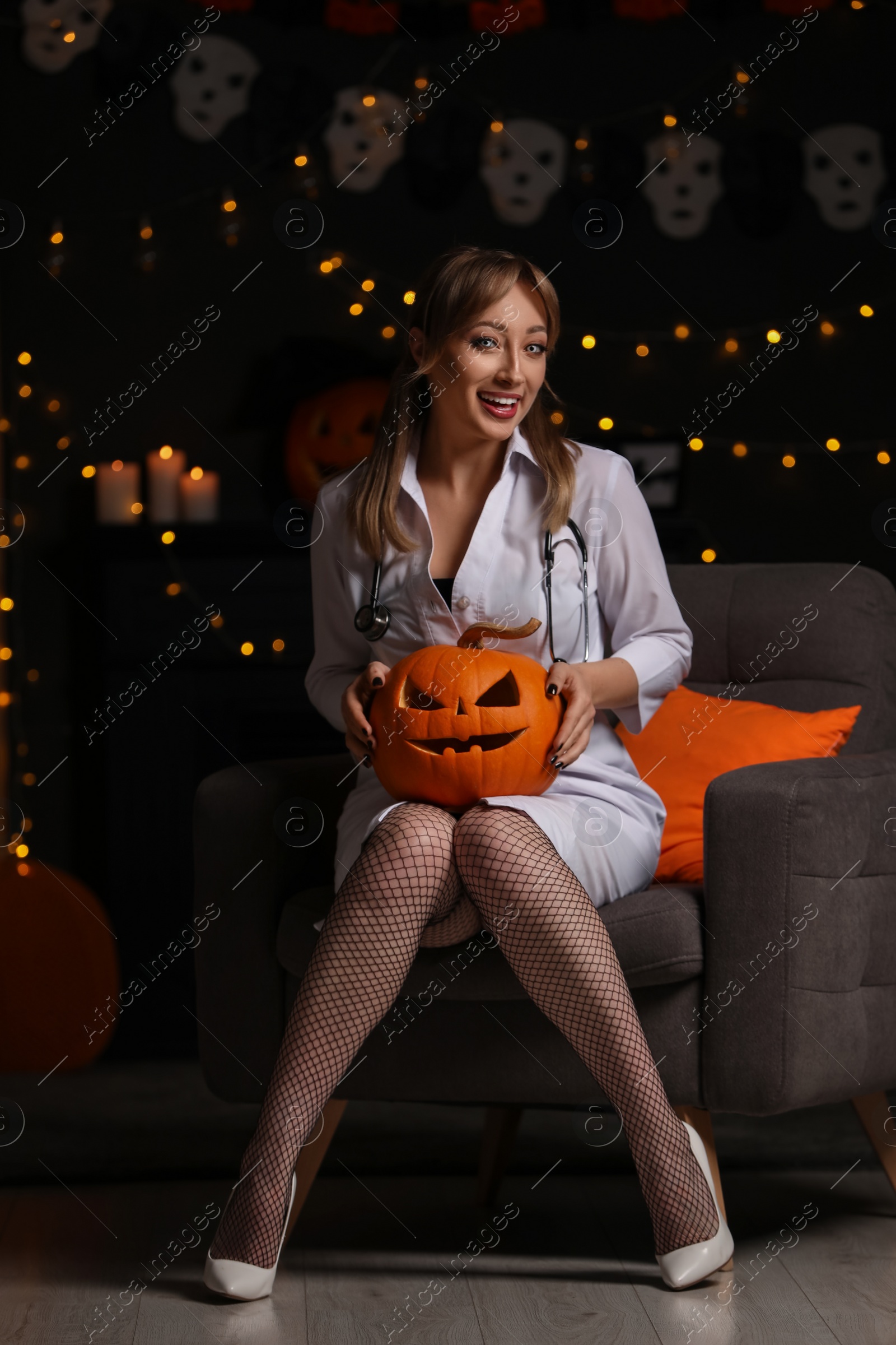 Photo of Woman in scary nurse costume with carved pumpkin against blurred lights indoors. Halloween celebration