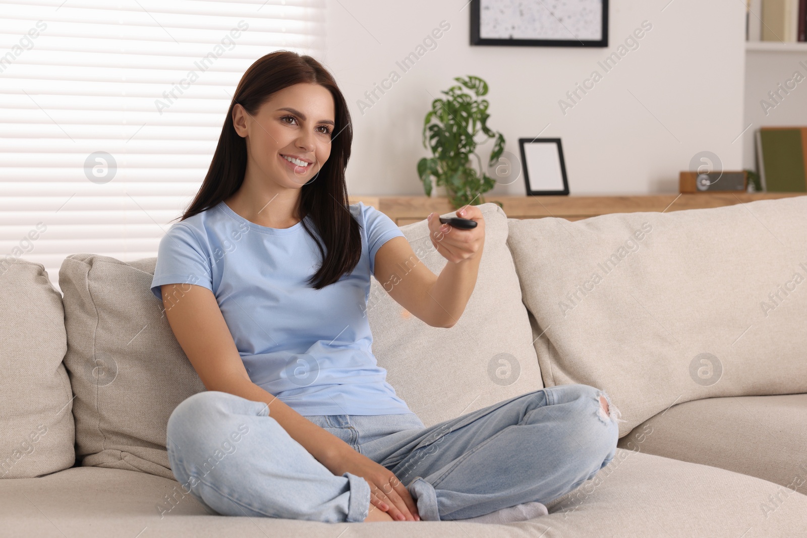 Photo of Happy woman changing TV channels with remote control on sofa at home