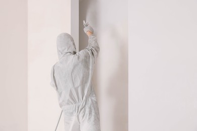 Photo of Decorator in uniform painting wall with sprayer indoors, back view