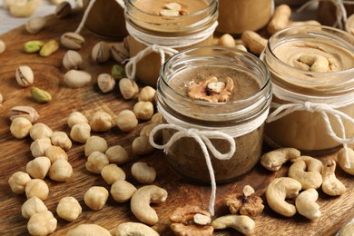 Photo of Tasty nut butters in jars and raw nuts on wooden board, closeup