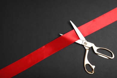 Photo of Stylish scissors and red ribbon on black background, flat lay with space for text. Ceremonial tape cutting