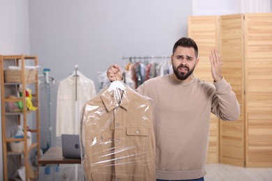 Photo of Dry-cleaning service. Emotional man holding hanger with jacket in plastic bag indoors