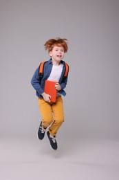 Photo of Happy schoolboy with backpack and book jumping on grey background
