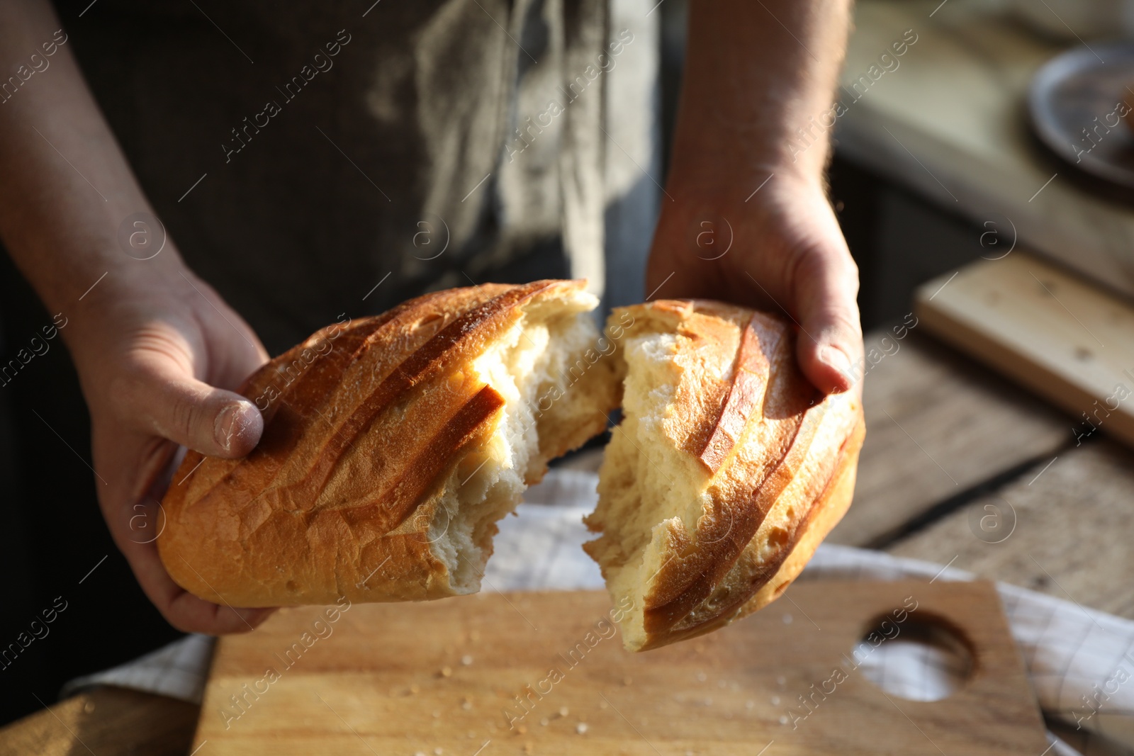 Photo of Man breaking loaf of fresh bread at wooden table indoors, closeup