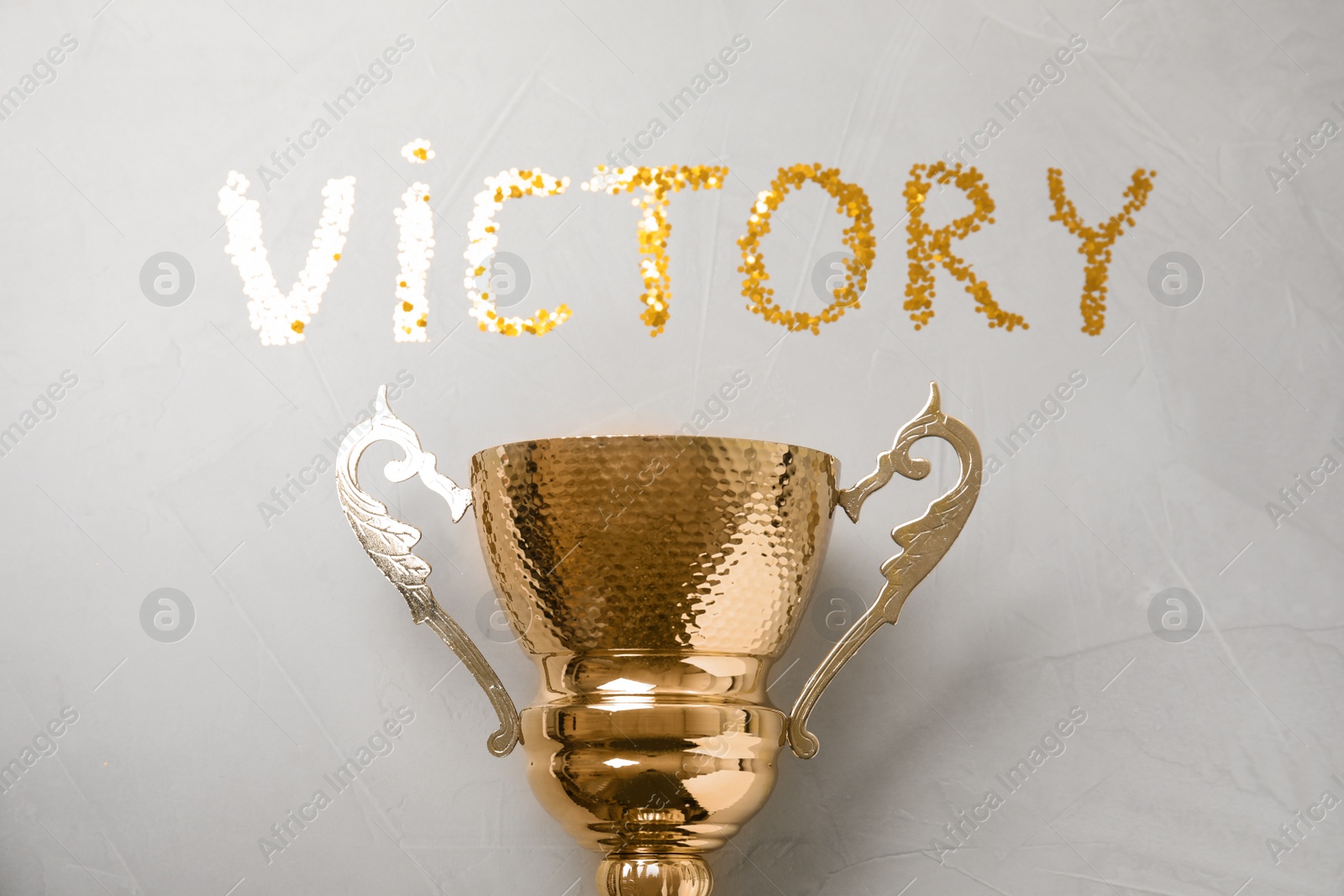 Photo of Gold trophy cup and word VICTORY made with confetti on light grey stone background, flat lay