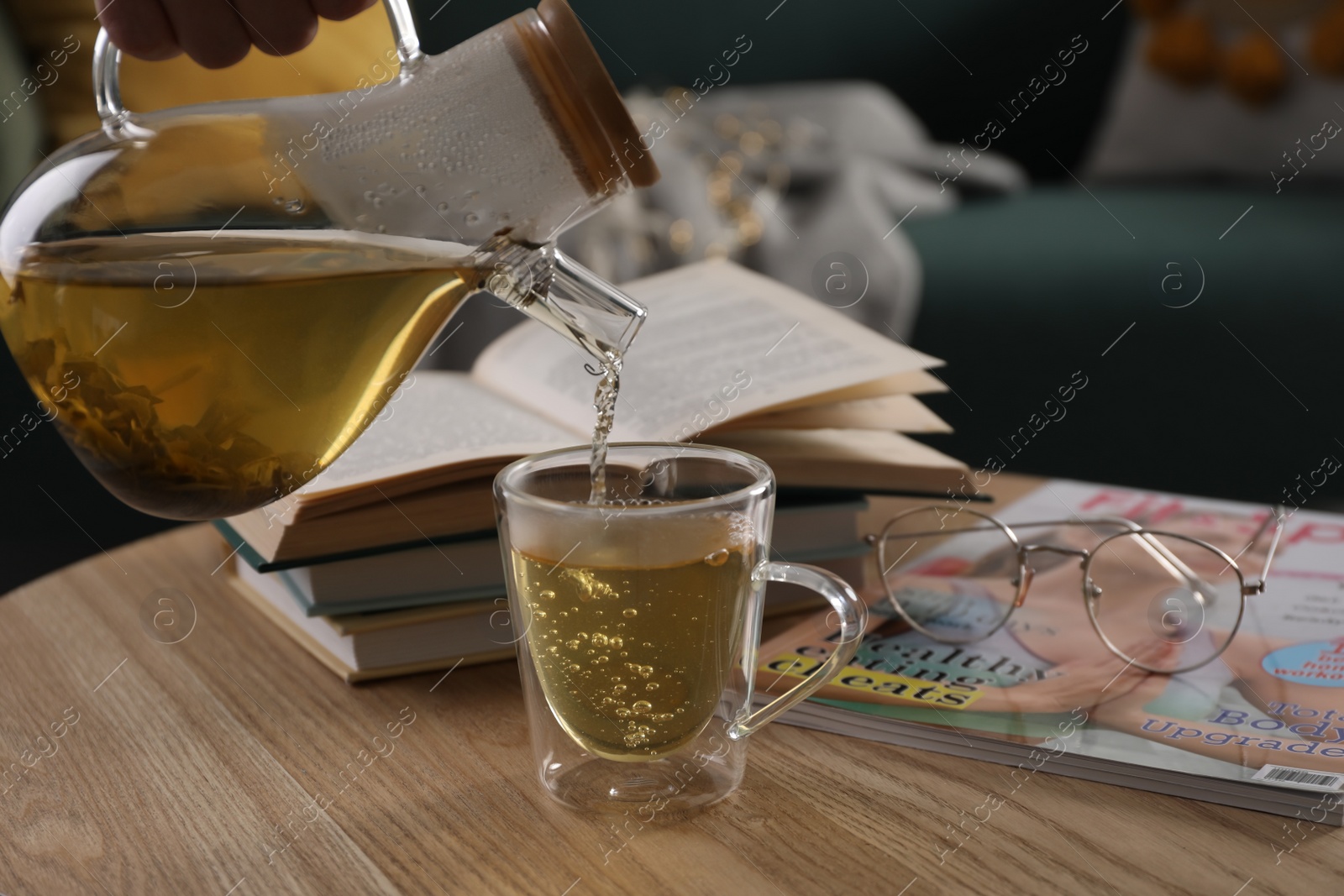 Photo of Woman pouring freshly brewed tea into cup at table in living room, closeup. Cozy home atmosphere