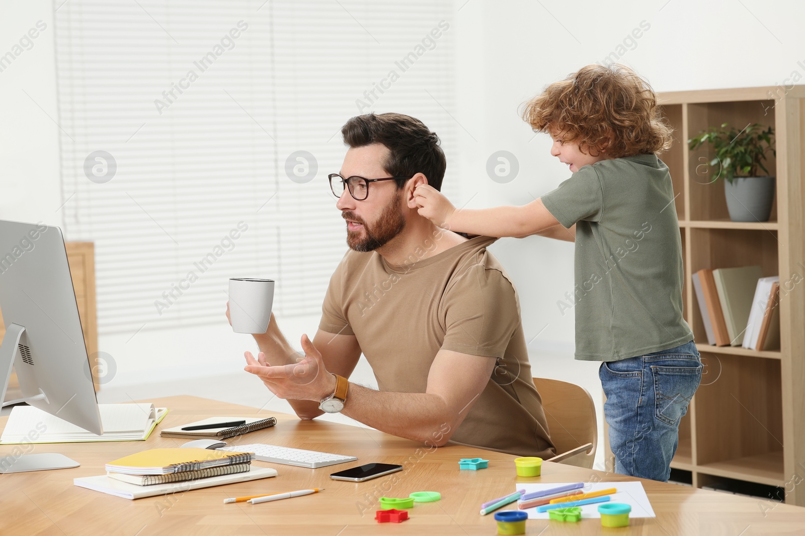 Photo of Little boy bothering his father at home. Man working remotely at desk