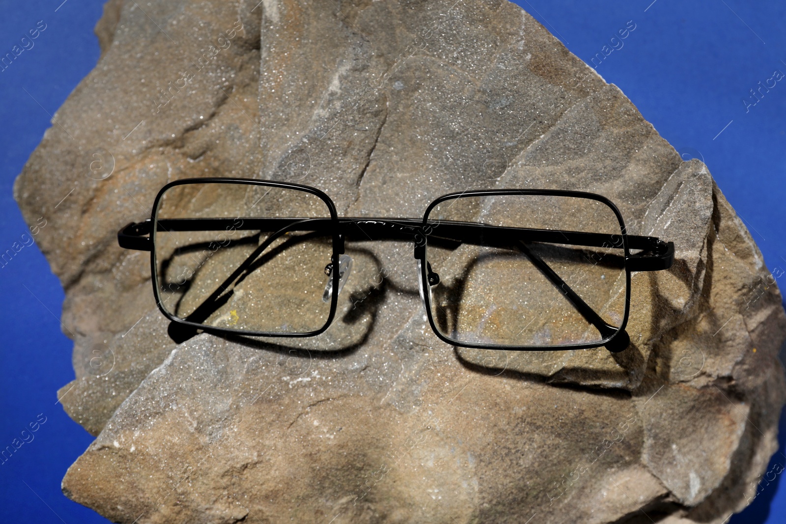 Photo of Stylish pair of glasses with black frame on stone against blue background, closeup