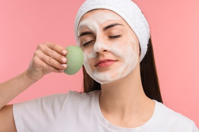 Young woman with headband washing her face using sponge on pink background