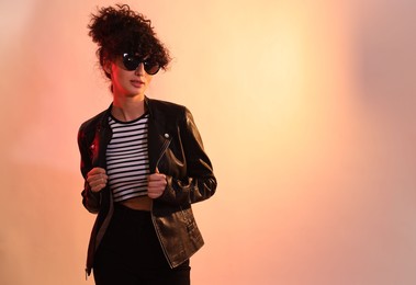 Photo of Beautiful young woman in black leather jacket and sunglasses posing on color background in neon lights. Space for text