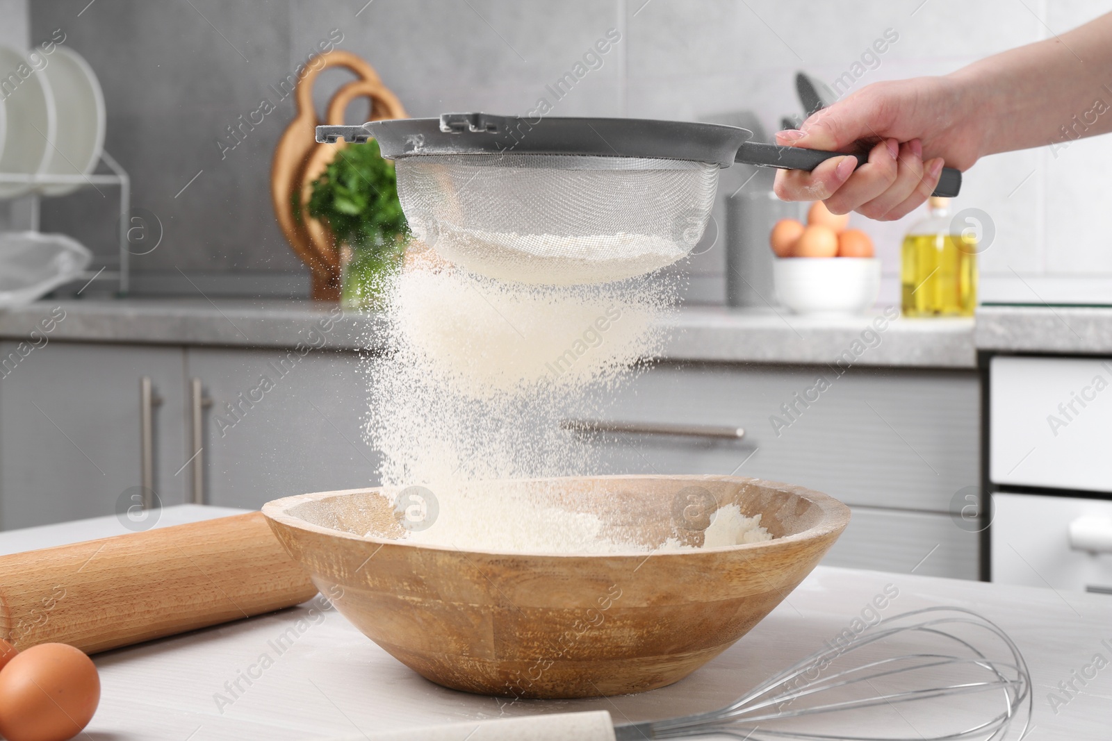 Photo of Woman sieving flour into bowl at white wooden table in kitchen, closeup