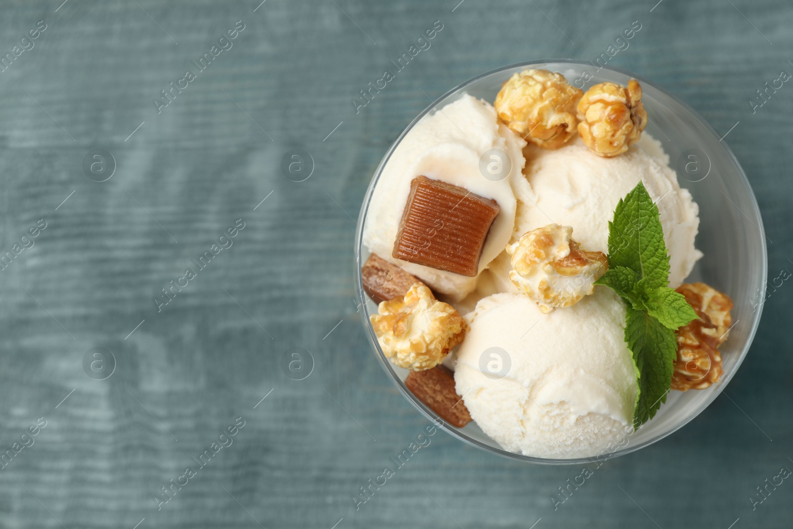 Photo of Delicious ice cream with caramel candies and popcorn in dessert bowl on blue wooden table, top view. Space for text