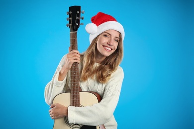 Photo of Young woman in Santa hat with acoustic guitar on light blue background. Christmas music