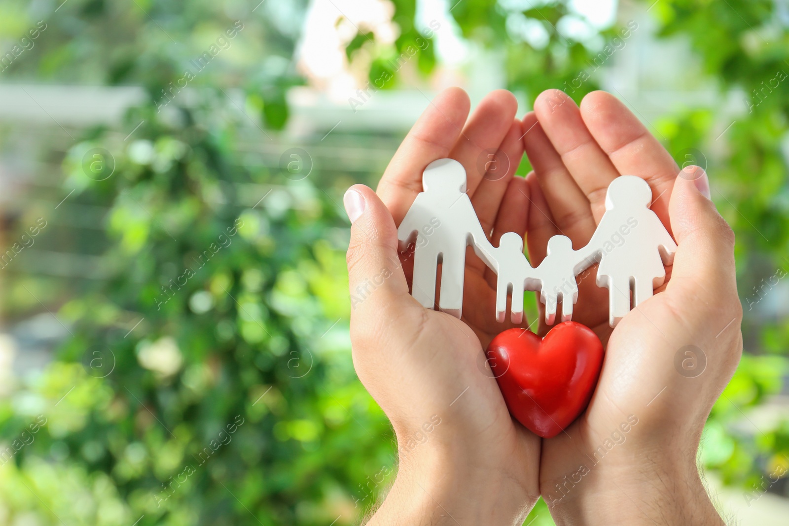 Photo of Young man holding wooden family figure and red heart in his hands against blurred background, closeup. Space for text