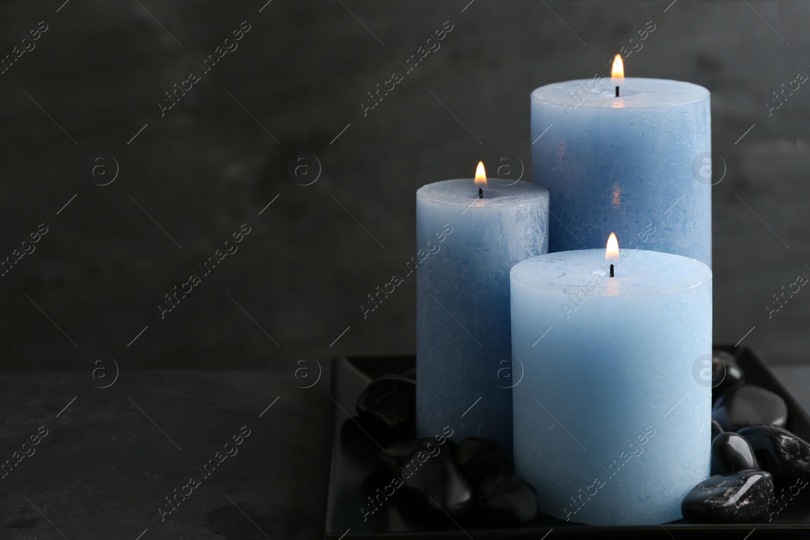 Photo of Dark plate with three burning candles and rocks on table, space for text