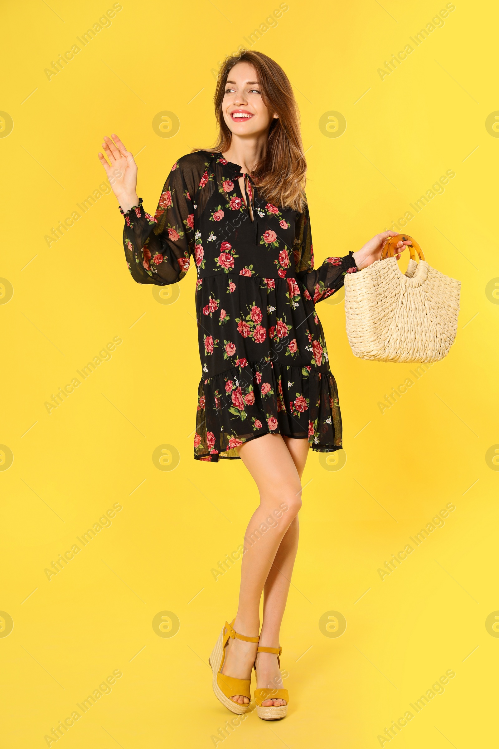 Photo of Young woman wearing floral print dress with straw bag on yellow background