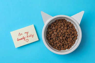 Photo of Feeding bowl of kibble with cat ears and cute note For My Lovely Kitty on light blue background, flat lay