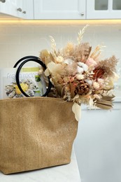 Photo of Stylish beach bag with beautiful bouquet of dried flowers and magazine on white table in kitchen