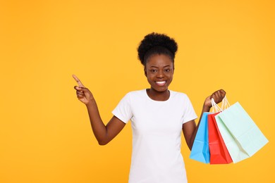 Happy young woman with shopping bags pointing at something on orange background. Space for text