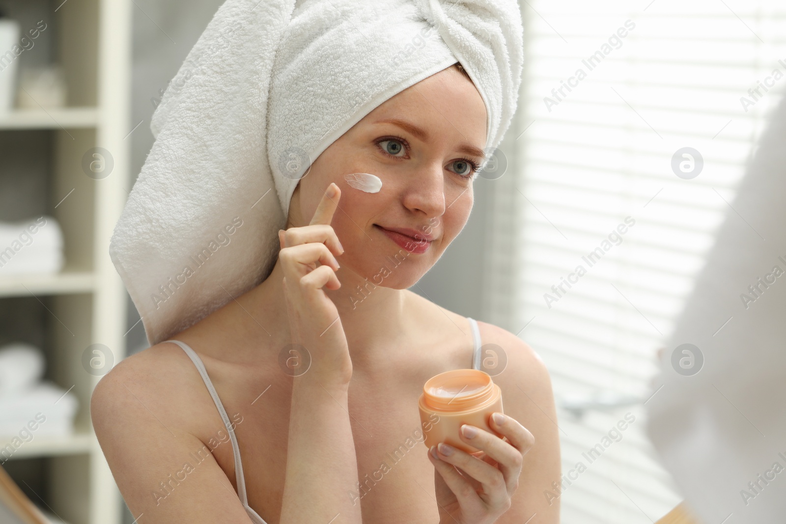 Photo of Beautiful woman with freckles applying cream onto her face near mirror in bathroom