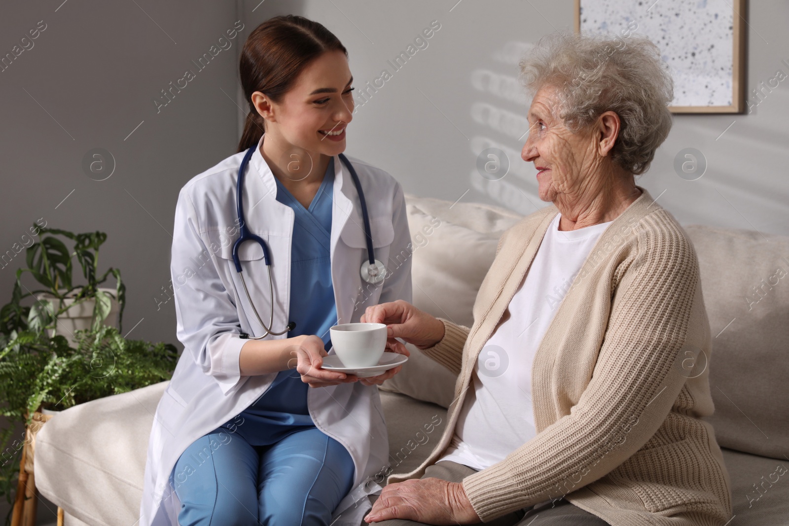 Photo of Young caregiver giving drink to senior woman in room Home health care service