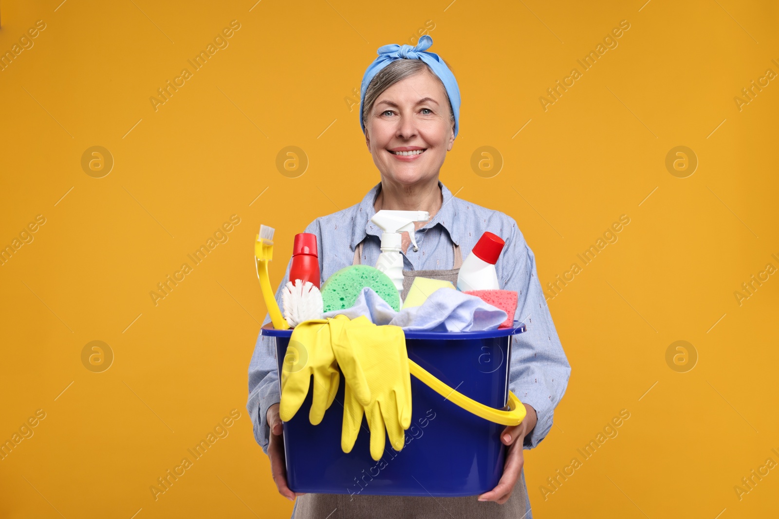 Photo of Happy housewife holding bucket with cleaning supplies on orange background