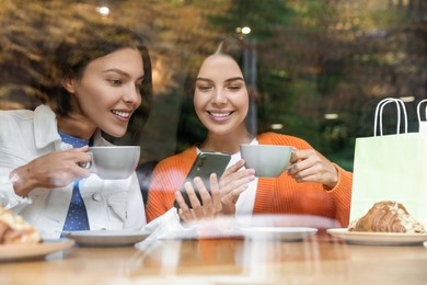 Photo of Special Promotion. Happy young women with cups of drinks using smartphone at table in cafe, view from outdoors