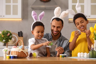 Photo of Happy African American father and his cute children painting Easter eggs at table in kitchen