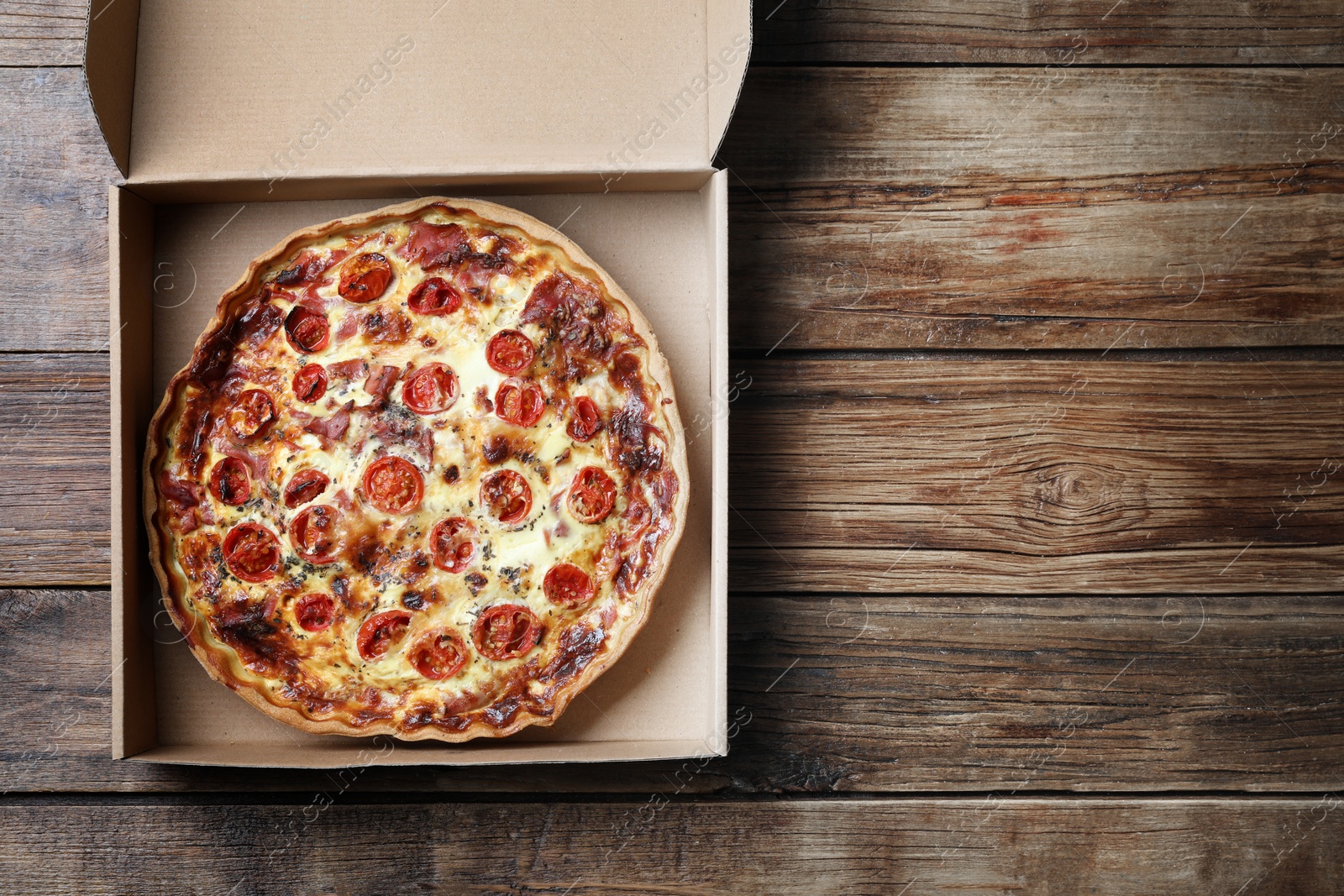 Photo of Delicious quiche with prosciutto and tomatoes in delivery box on wooden table, top view. Space for text