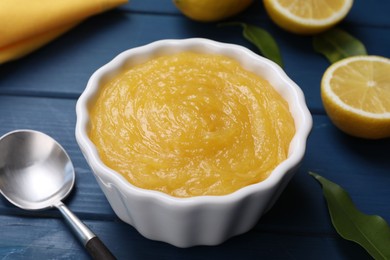 Photo of Delicious lemon curd in bowl, fresh citrus fruits and spoon on blue wooden table, closeup