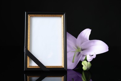 Image of Funeral photo frame with ribbon and violet lily on table against black background