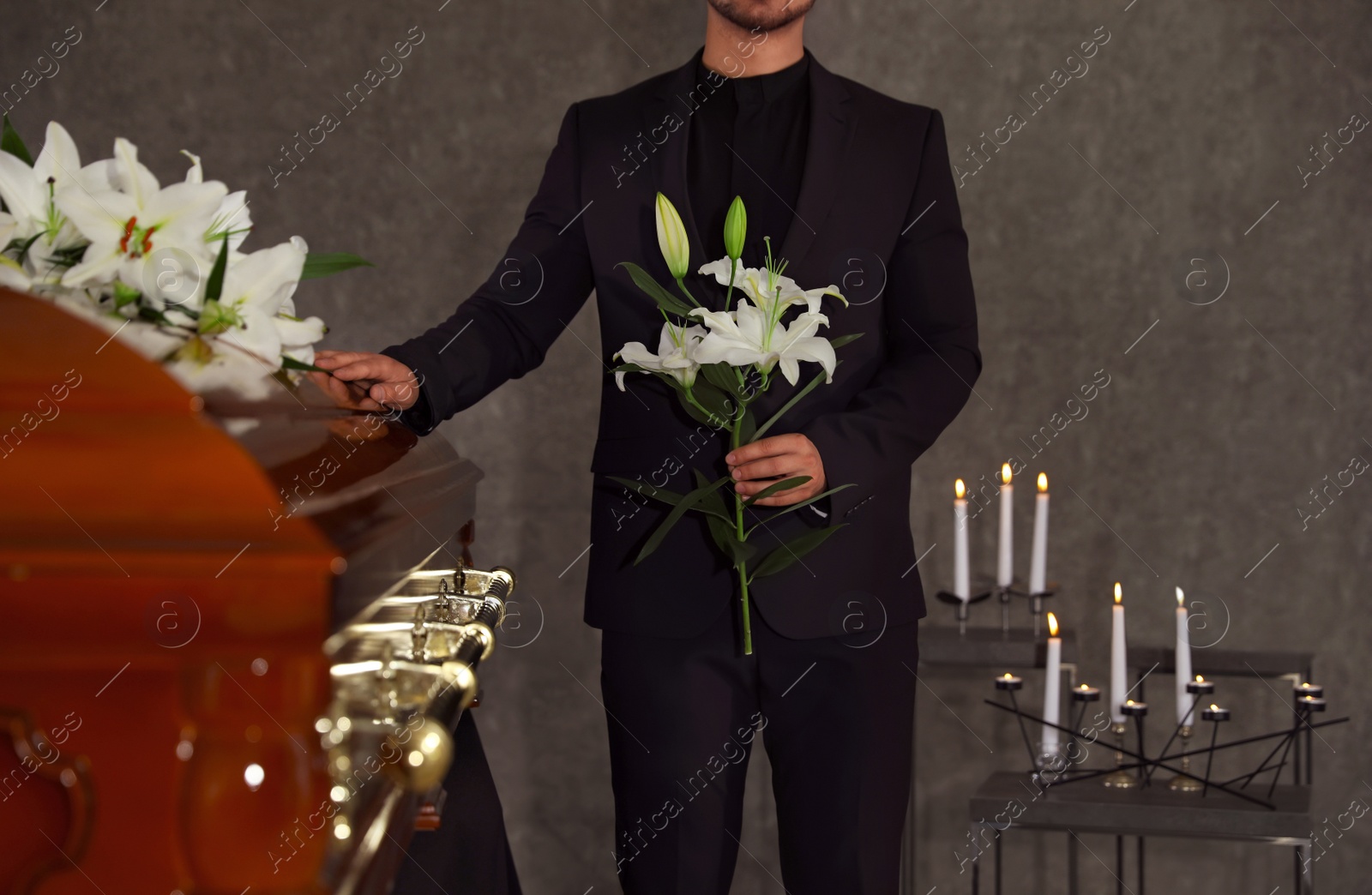 Photo of Young man with white lilies near casket in funeral home, closeup