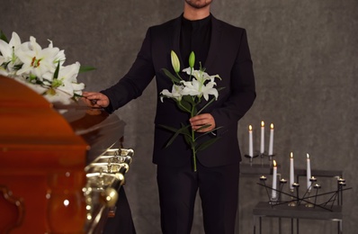 Photo of Young man with white lilies near casket in funeral home, closeup