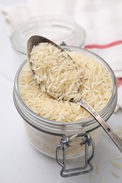 Photo of Glass jar and spoon with raw rice on white table, closeup