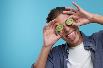 Photo of Smiling man covering eyes with halves of kiwi and showing tongue on light blue background. Space for text