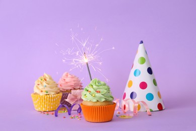 Photo of Birthday cupcakes with burning sparkler, party hat and gift box on violet background