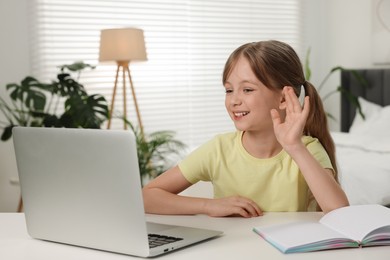 Photo of Cute girl waving hello during online lesson via laptop at white table indoors