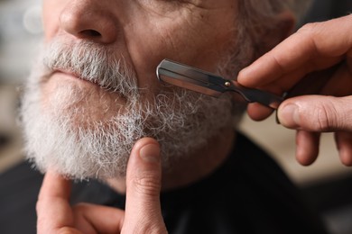 Photo of Professional barber shaving client's beard with blade in barbershop, closeup