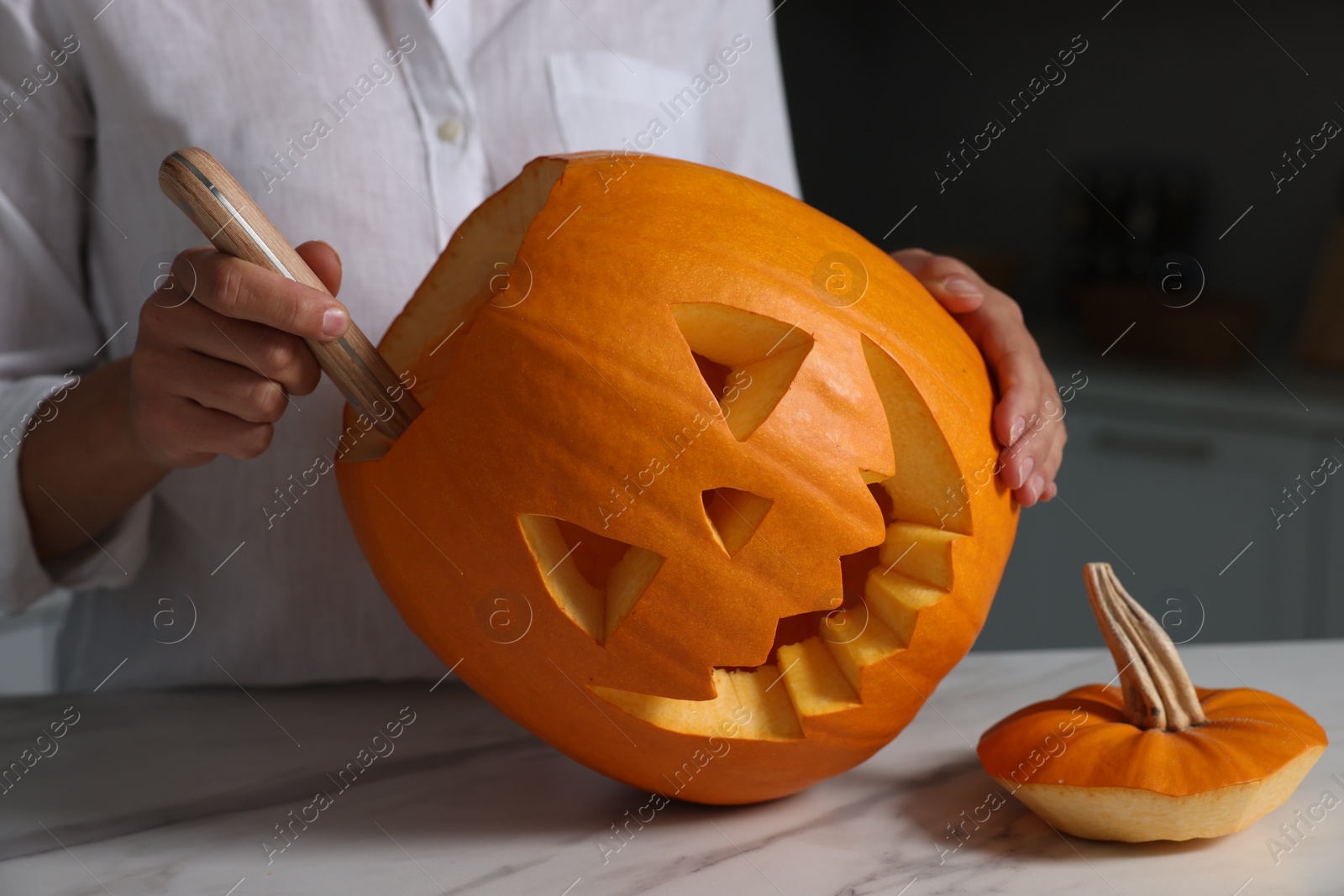 Photo of Woman carving pumpkin for Halloween at white marble table in kitchen, closeup