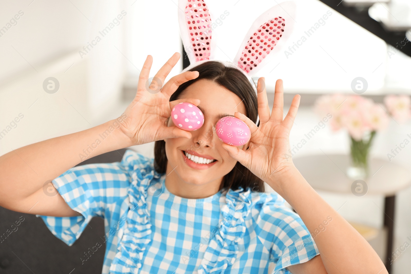 Photo of Beautiful woman in bunny ears headband holding Easter eggs near eyes at home