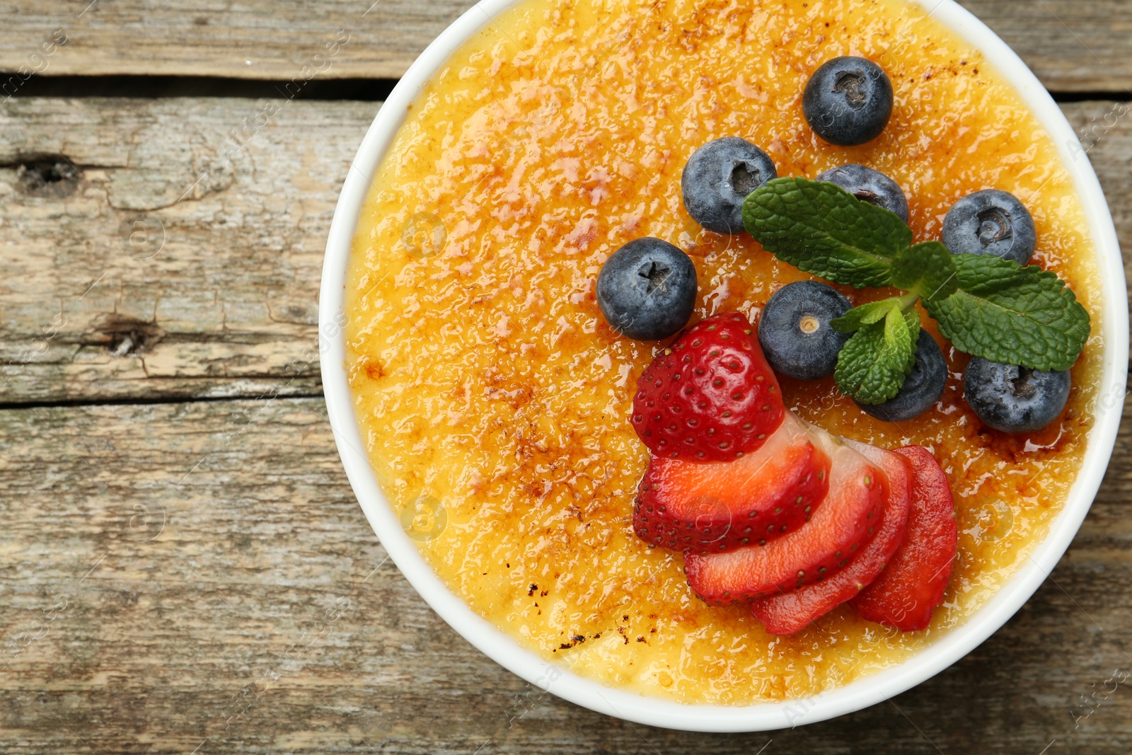 Photo of Delicious creme brulee with berries and mint in bowl on wooden table, top view. Space for text