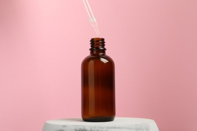 Photo of Dripping face serum into bottle on marble stand against pink background, closeup