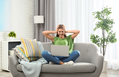 Emotional young woman with laptop celebrating victory on sofa at home