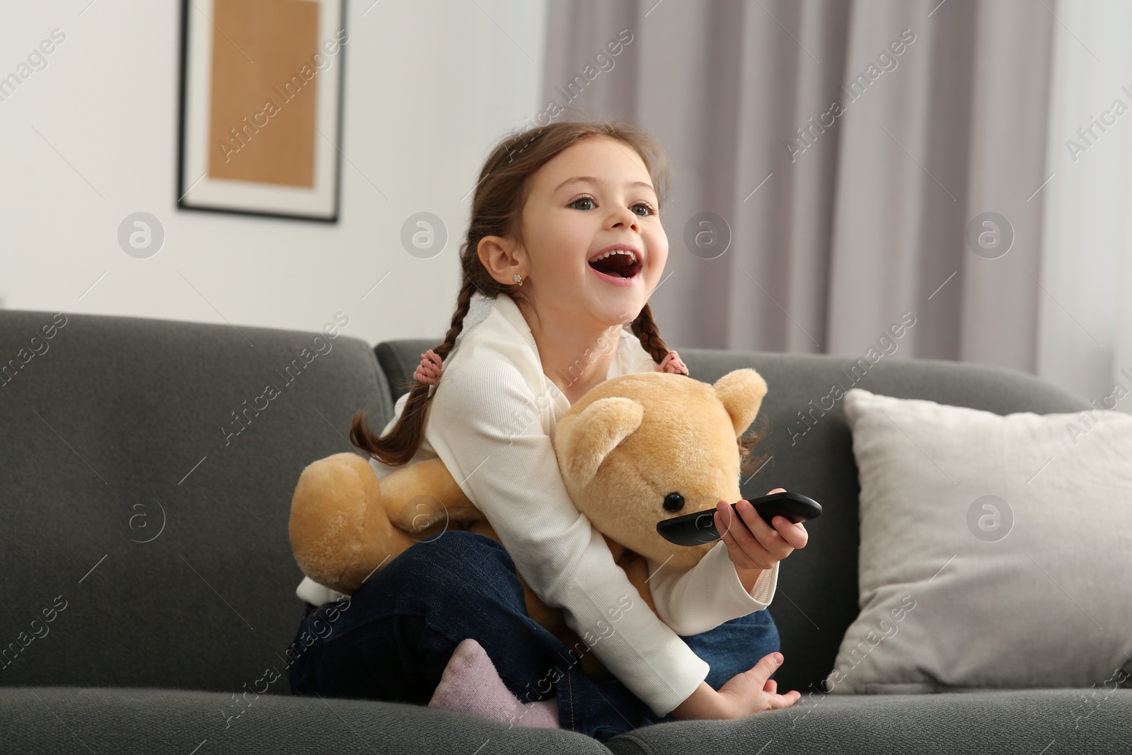 Photo of Happy girl changing TV channels with remote control on sofa at home