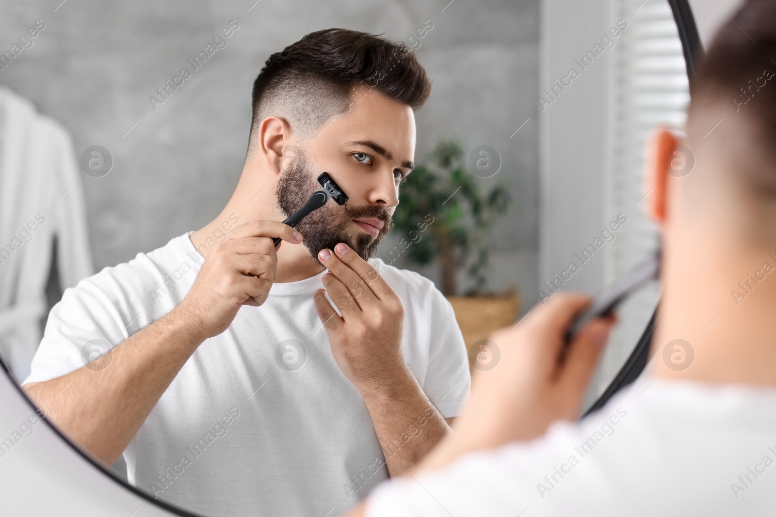 Photo of Handsome young man shaving with razor near mirror in bathroom