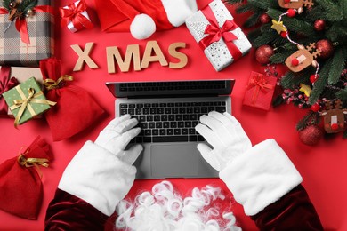 Photo of Santa Claus using laptop, closeup. Gift boxes and Christmas decor on red background, top view