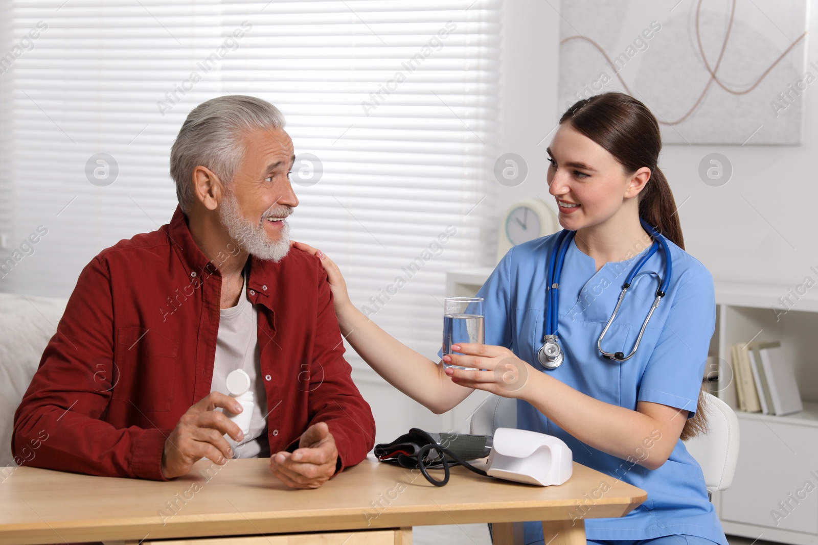 Photo of Young healthcare worker giving glass of water to senior man at wooden table indoors